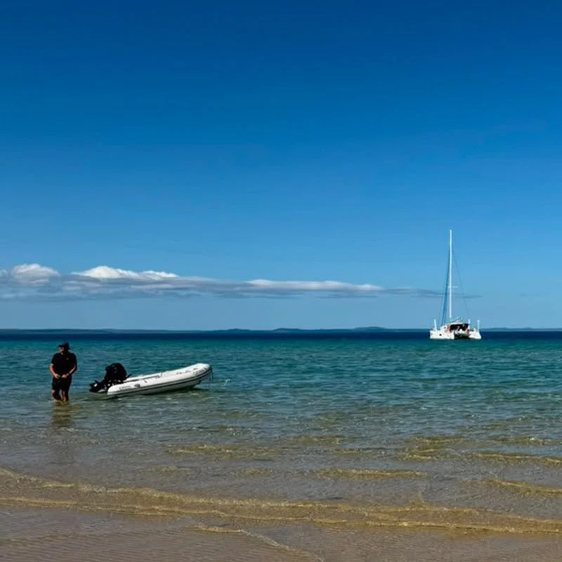 Inflatable tender at a beach with a sailing catamaran offshore.