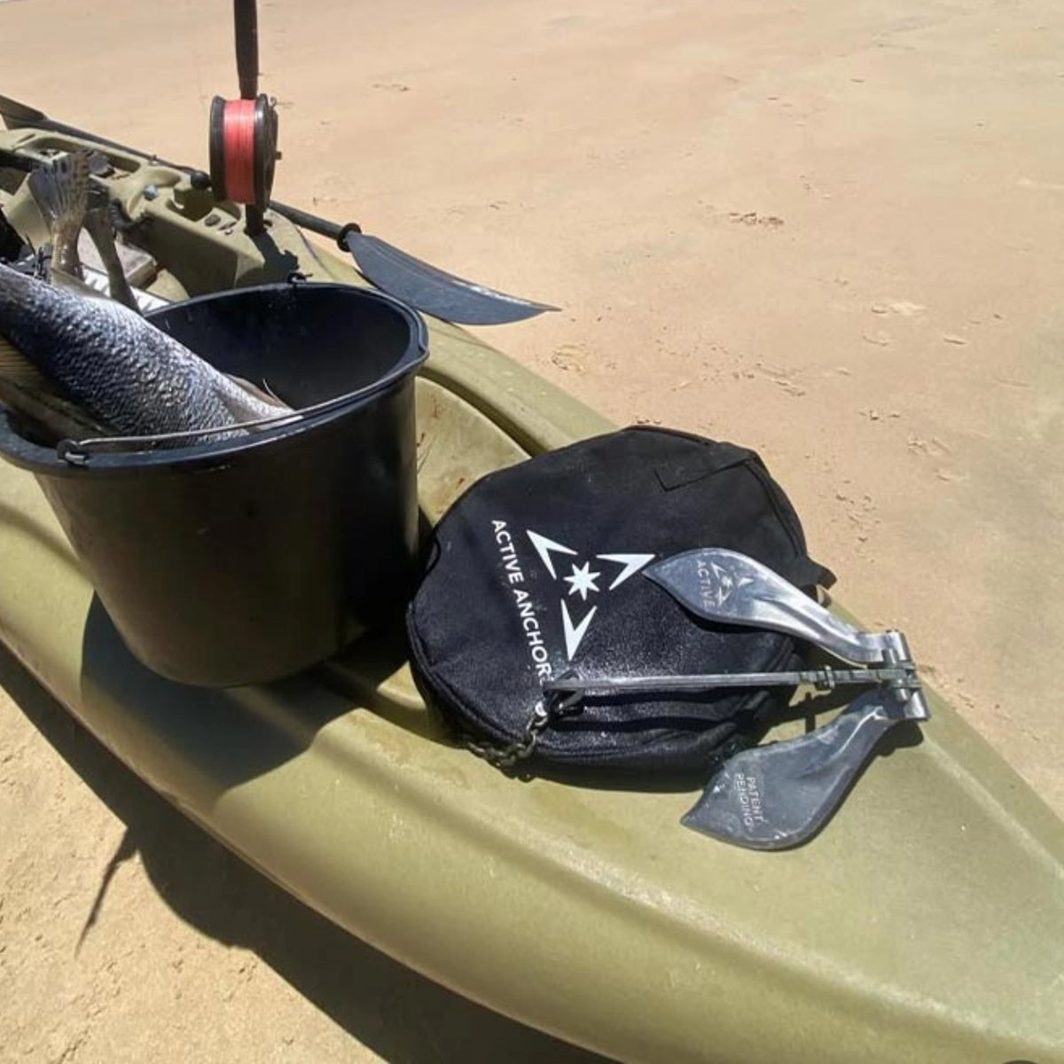 Active Anchors DF200 boat anchor resting on its black carry bag alongside fishing equipment in a kayak on a sandy beach.